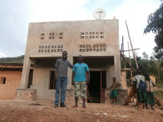 Claude in a blue t-shirt posing in front of his commercial house in Bucumbi village 